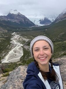 A person in a beanie and jacket smiles for a selfie on a rocky ledge with braided river, green valley, and snow-capped mountains in the background.