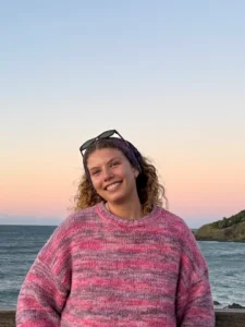 A person wearing a pink striped sweater and sunglasses on their head smiles while standing in front of the ocean at sunset.