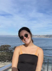 A woman wearing sunglasses and a black top smiles while standing in front of a rocky shoreline and ocean under a partly cloudy sky.