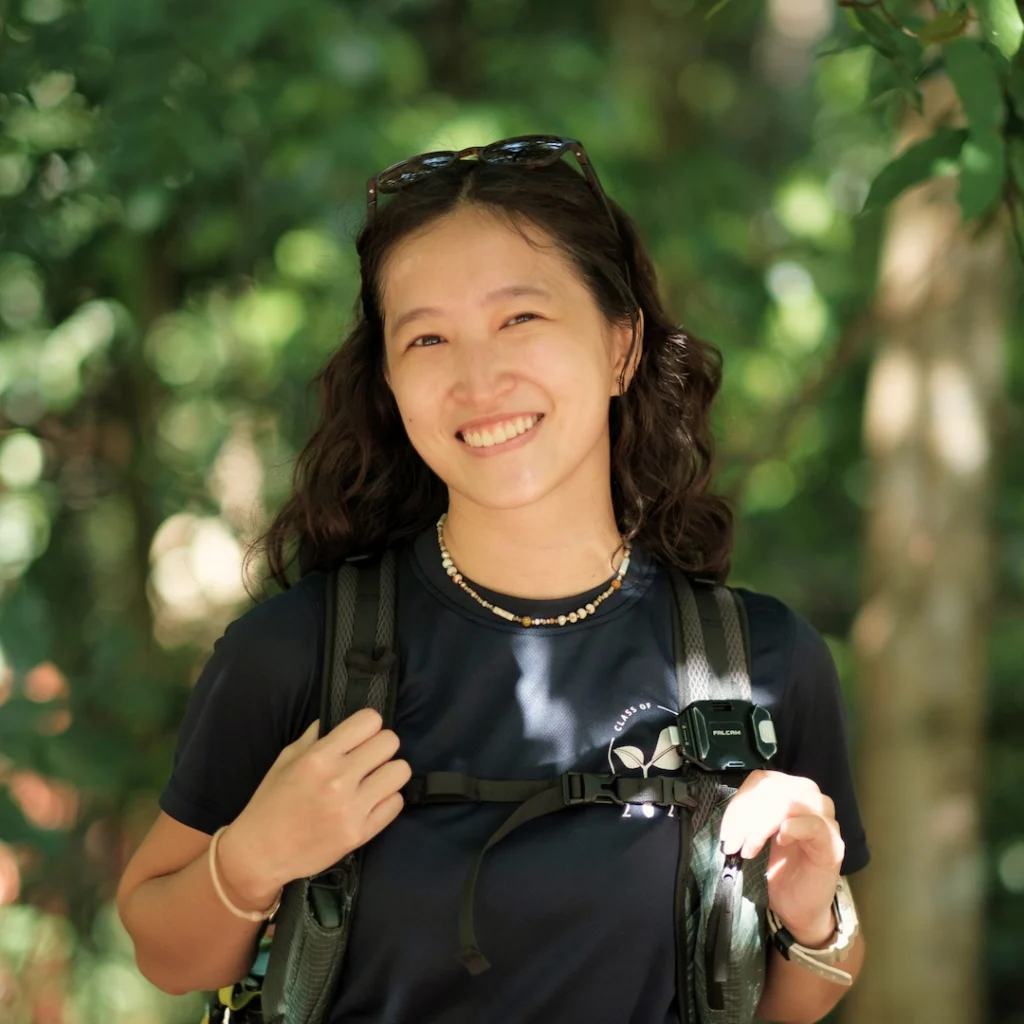 A person with long curly hair, wearing a dark t-shirt and backpack, smiles outdoors in a sunlit, leafy area.